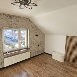 Patryk Chyrzynski - Pokój na poddaszu po remoncie: dekoracyjna ściana z efektem betonu, drewniana podłoga, panel z drewnianych listew i biała pufa. Okno z widokiem na ulicę.