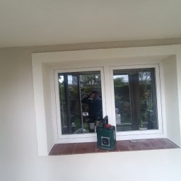 Patryk Chyrzynski - Wykończone szpachlowaniem ściany wokół okna z parapetem wyłożonym brązowymi płytkami. Widoczne narzędzia i odbicie osoby w szybie okiennej.