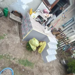 Wykończone schody wejściowe z szarymi płytkami, z widocznymi elementami mocującymi. Na górze tarasu pies leży na krześle, obok stoi osoba. Prace w toku.