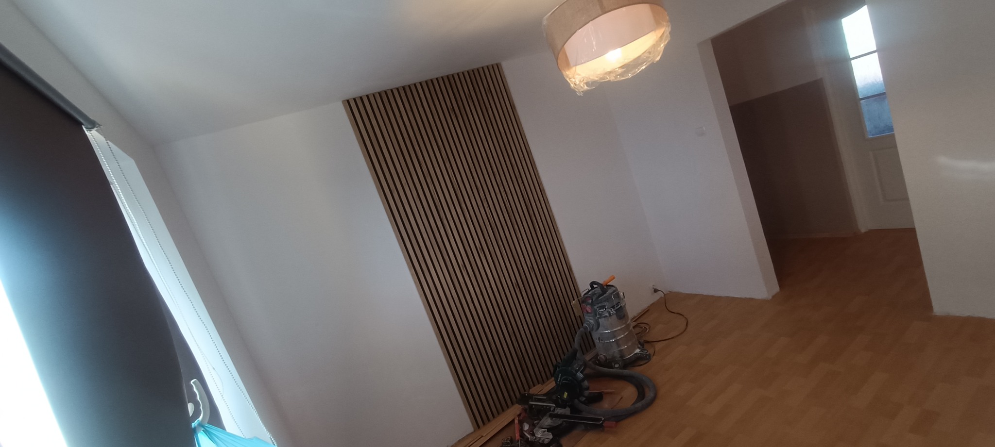 Wnętrze z panelami ściennymi z drewna, widoczne narzędzia i odkurzacz. Na suficie lampa w folii ochronnej. Podłoga wyłożona panelami. W tle widoczne drzwi.