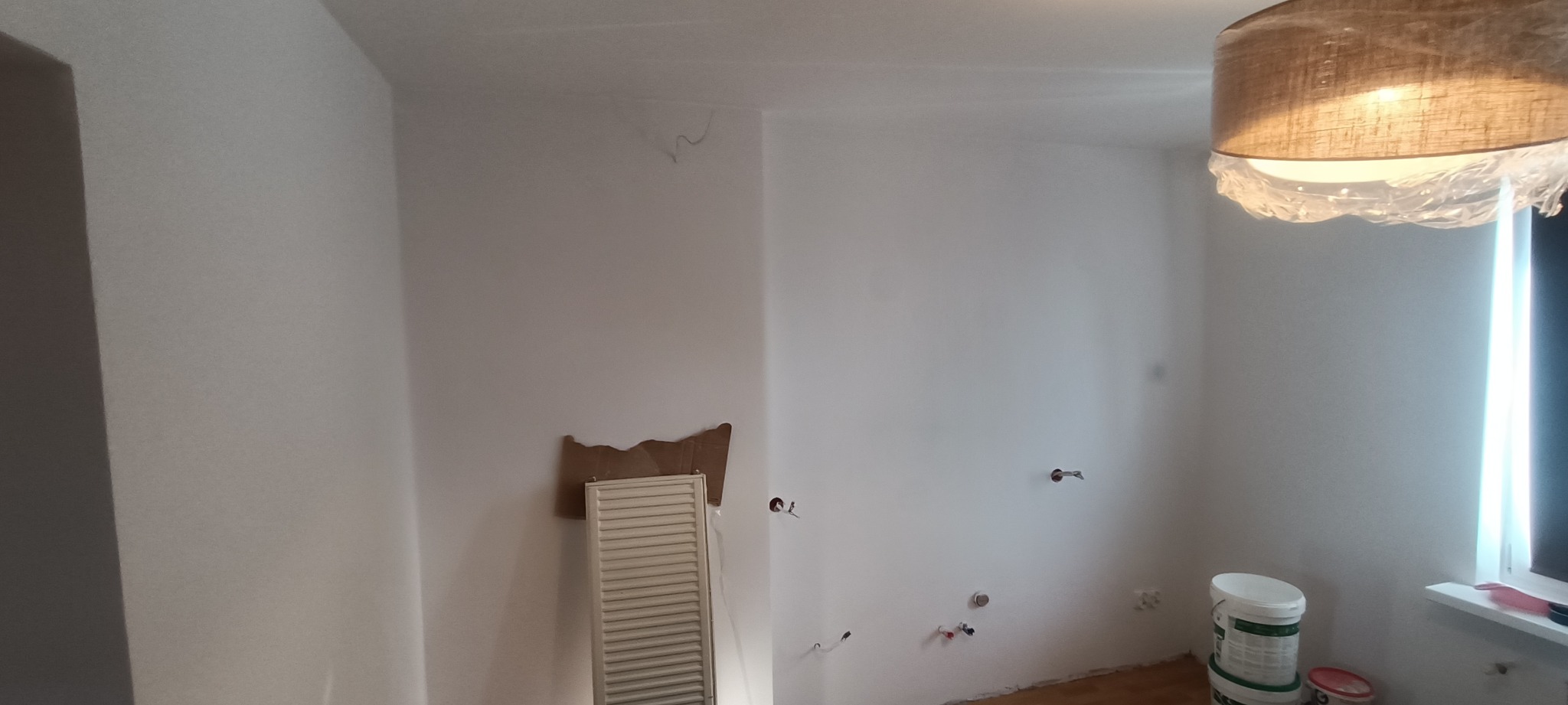 Białe ściany w pokoju po malowaniu, widoczne puszki z farbą, zakryta lampa. Przy ścianie zabezpieczony grzejnik. Minimalistyczne wnętrze przed wykończeniem.