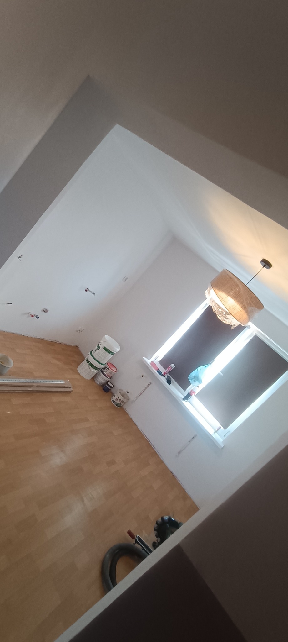 Pokój po remoncie: drewniana podłoga, białe ściany, okno z roletami, lampa wisząca w folii. Widoczne farby i narzędzia malarskie.