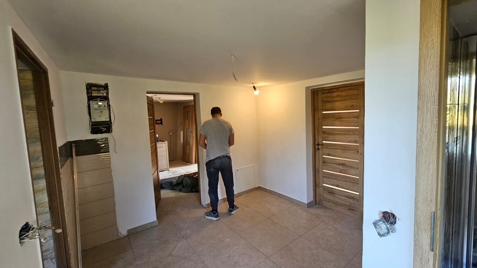 Wnętrze po odświeżeniu: białe ściany, beżowa podłoga, drewniane drzwi z przeszkleniami. Widoczny elektryk przy pracy, odsłonięty licznik i instalacja elektryczna.