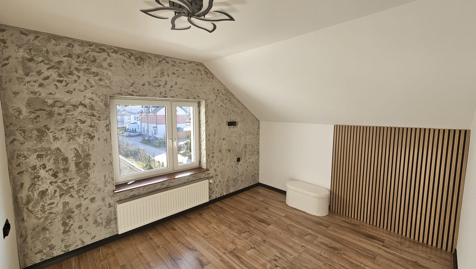 Pokój na poddaszu po remoncie: dekoracyjna ściana z efektem betonu, drewniana podłoga, panel z drewnianych listew i biała pufa. Okno z widokiem na ulicę.