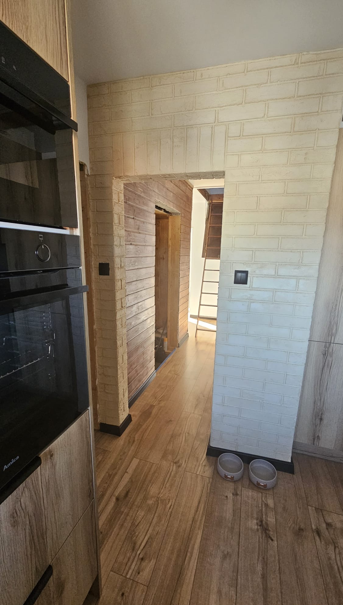 Wnętrze domu z drewnianą podłogą i ścianami wykończonymi imitacją cegły w odcieniach beżu i bieli. Widoczny fragment kuchni i przejście do innego pomieszczenia ze schodami.