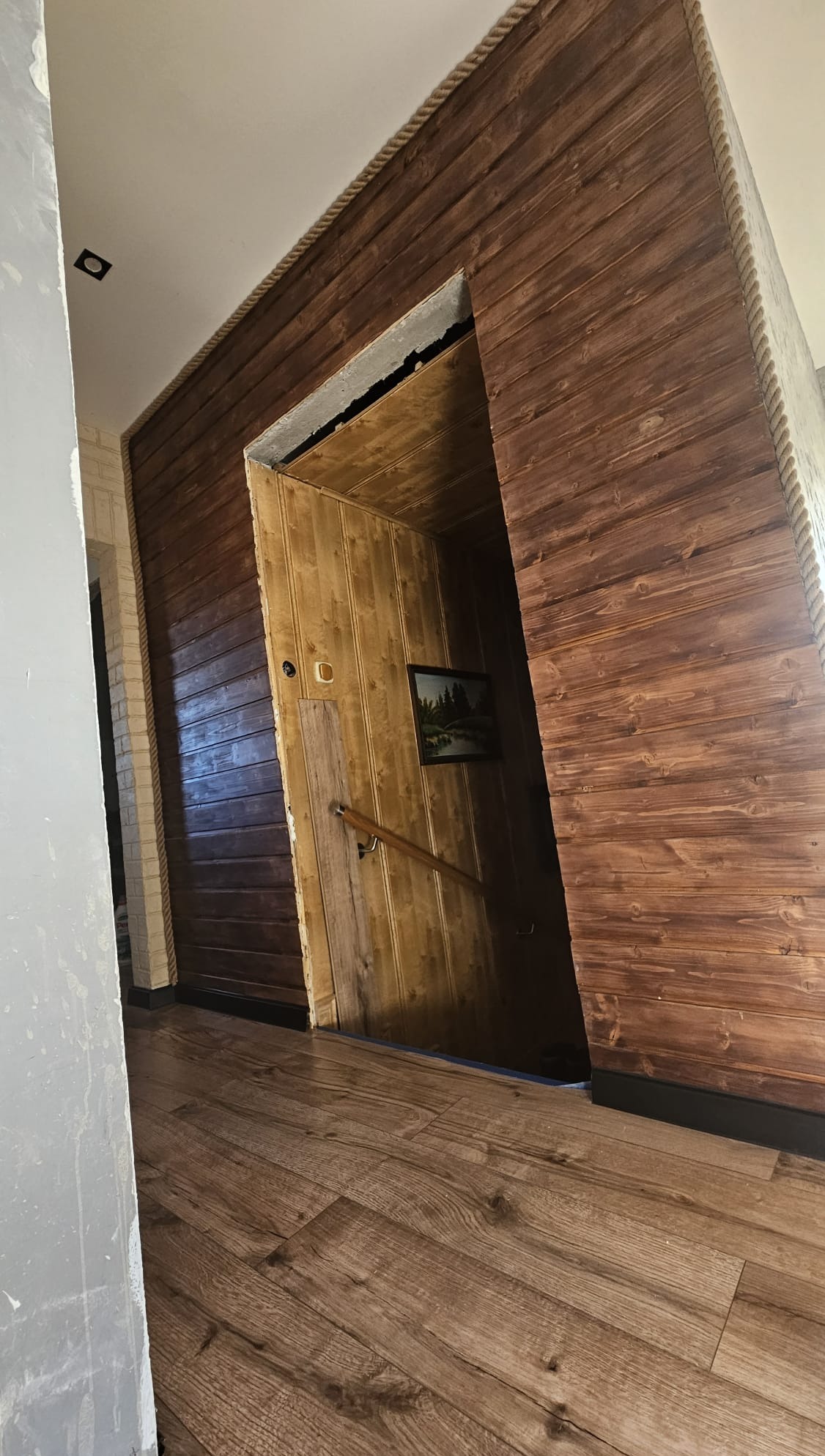 Wykończenie wnętrza z drewnianą boazerią w ciepłych odcieniach brązu, zdobione grubym sznurem. Widoczne drewniane schody i podłoga z paneli.