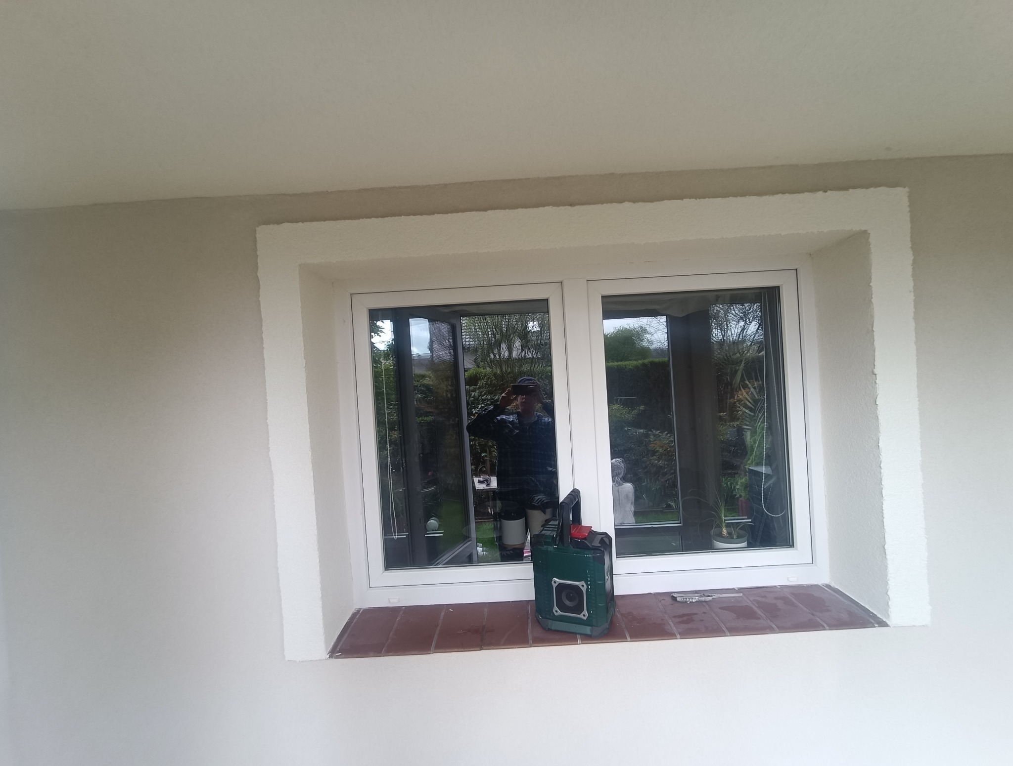 Wykończone szpachlowaniem ściany wokół okna z parapetem wyłożonym brązowymi płytkami. Widoczne narzędzia i odbicie osoby w szybie okiennej.