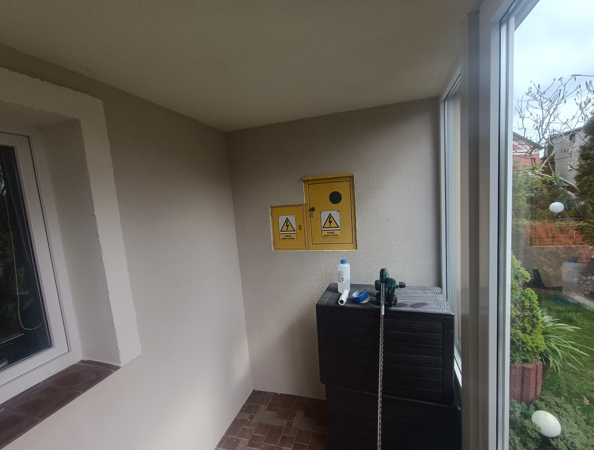 Wnętrze balkonu po remoncie: świeżo pomalowane ściany, żółte skrzynki elektryczne i narzędzia na czarnej skrzyni. Widok na zieleń przez okno.