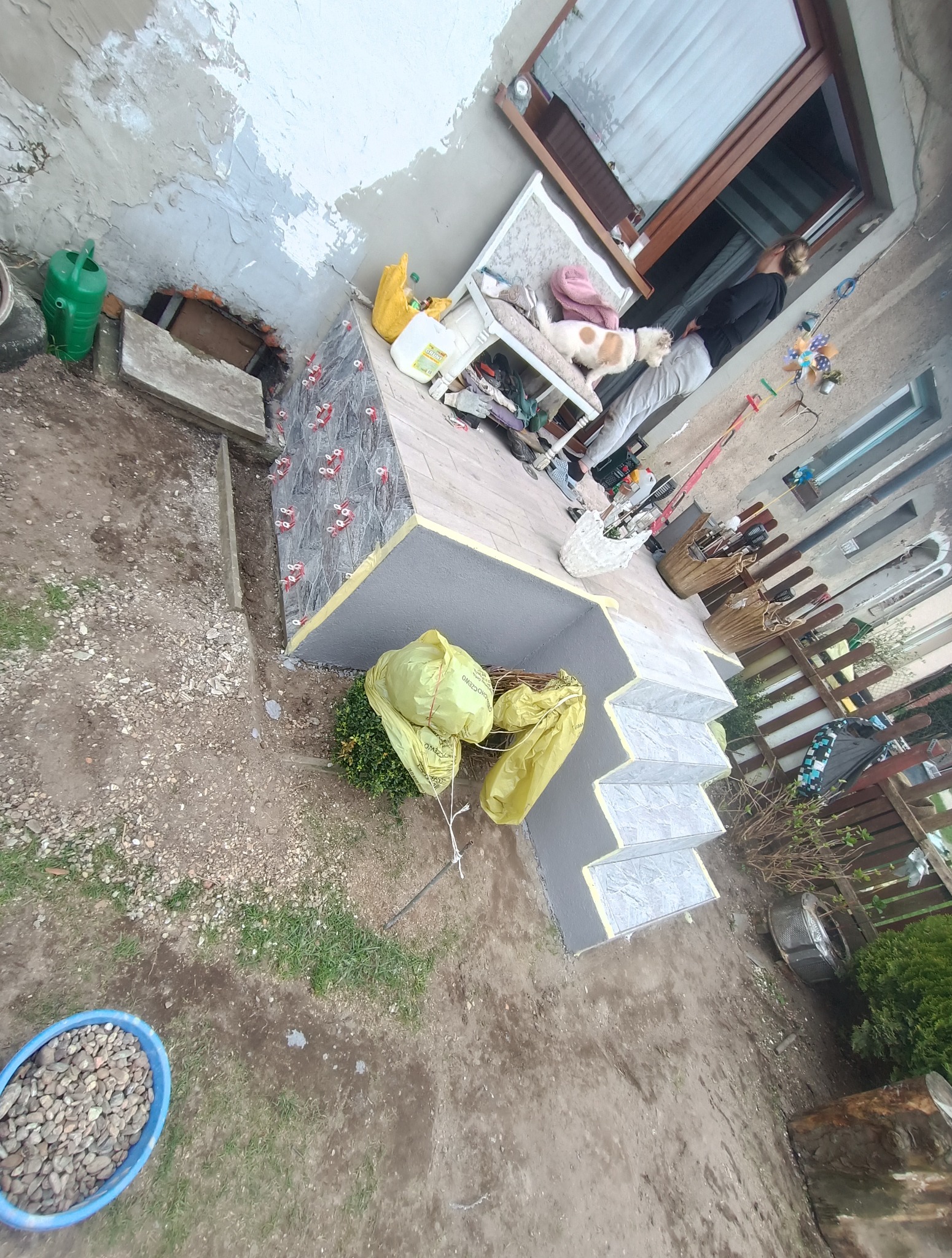 Wykończone schody wejściowe z szarymi płytkami, z widocznymi elementami mocującymi. Na górze tarasu pies leży na krześle, obok stoi osoba. Prace w toku.