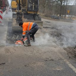 Daniel Szajc - Pracownik w pomarańczowej kamizelce tnie asfaltową nawierzchnię drogi za pomocą piły spalinowej, obok stoi żółta koparka, w tle drzewa i maszt telekomunikacyjny.