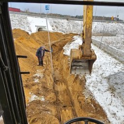 Daniel Szajc - Widok z kabiny koparki na wykopany rów w ziemi, pracownik mierzy głębokość, w tle ośnieżone pobocze drogi.