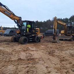 Daniel Szajc - Dwa żółte koparki kołowe SANY i CAT na placu budowy, teren przygotowany, widoczne ślady opon i łyżki, w tle las i samochody dostawcze.