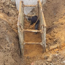 Daniel Szajc - Pracownik z łopatą w wykopie zabezpieczonym metalową szalunką, obok betonowy zbiornik szamba.