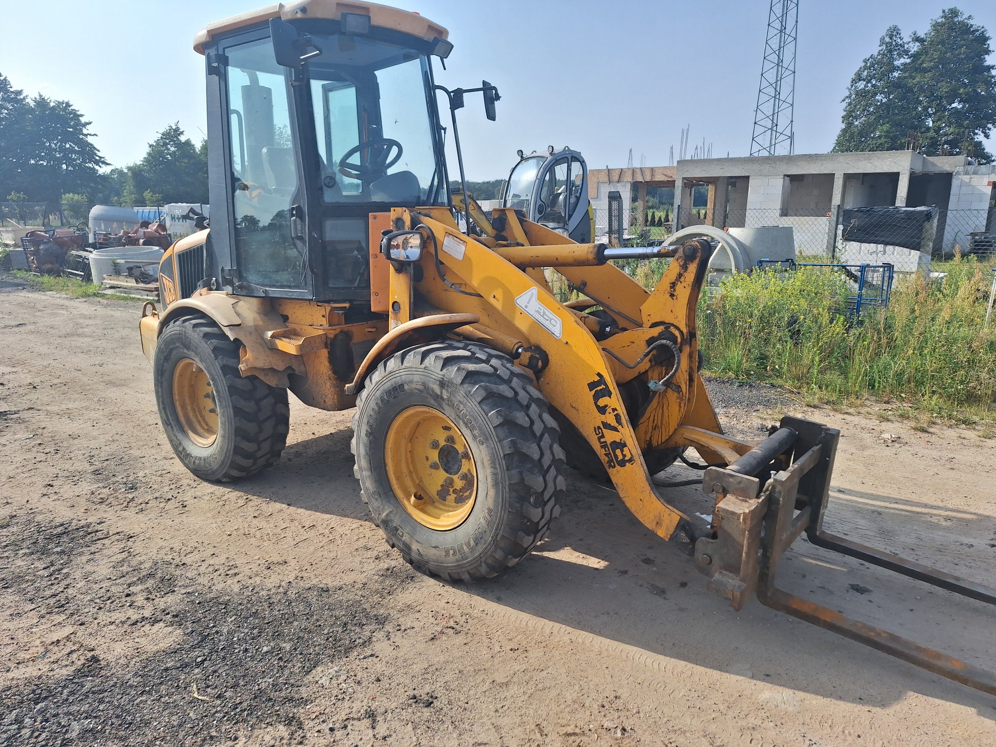 Żółta ładowarka teleskopowa JCB z widłami, stojąca na ziemi, w tle budowa domu w stanie surowym i słup wysokiego napięcia.