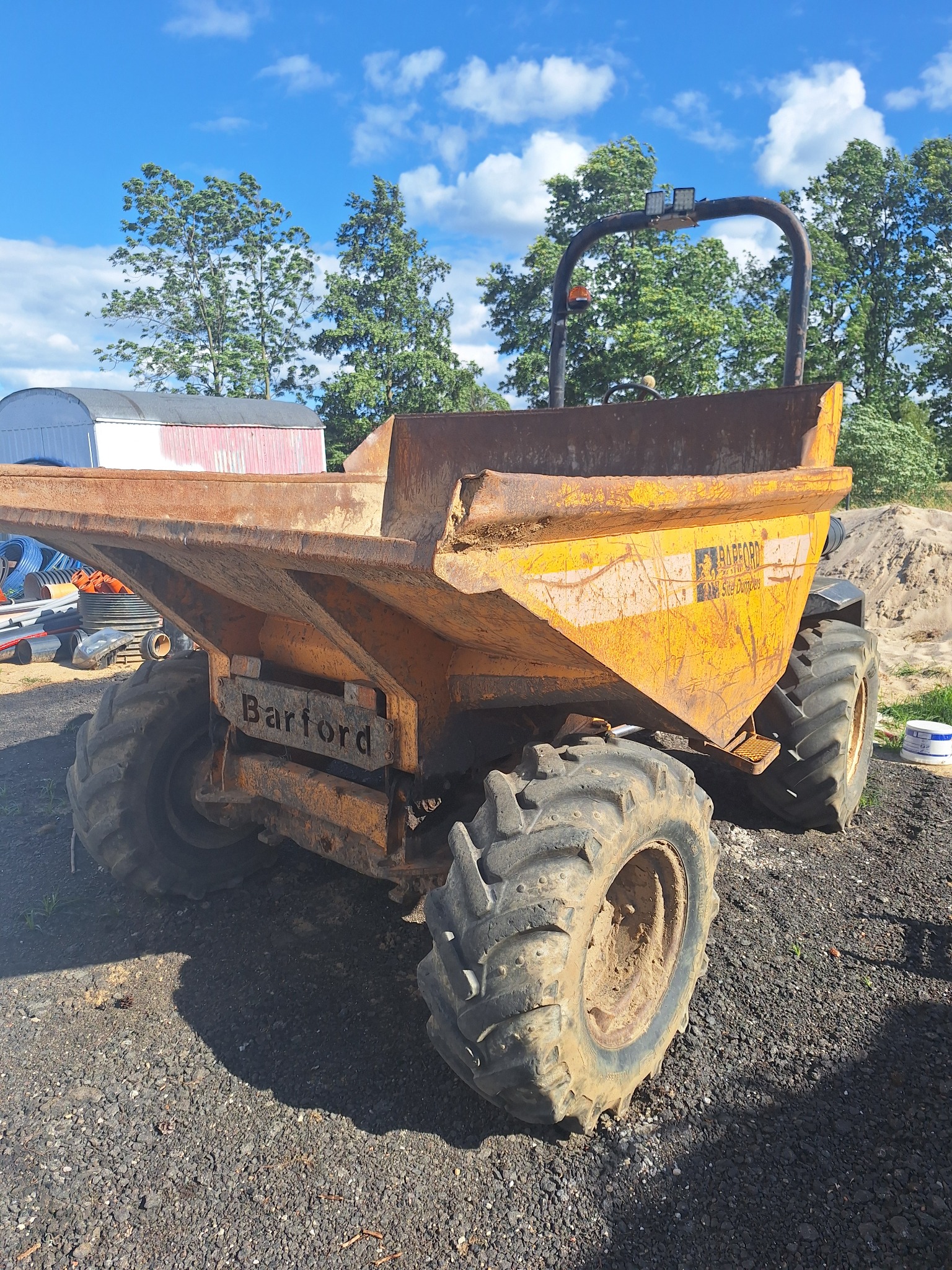 Używana, żółta wywrotka budowlana Barford Site Dumper z widocznymi śladami rdzy i zużycia, stojąca na żwirowym podłożu, z stertą piasku w tle i budynkiem gospodarczym w oddali, sfotografowana...