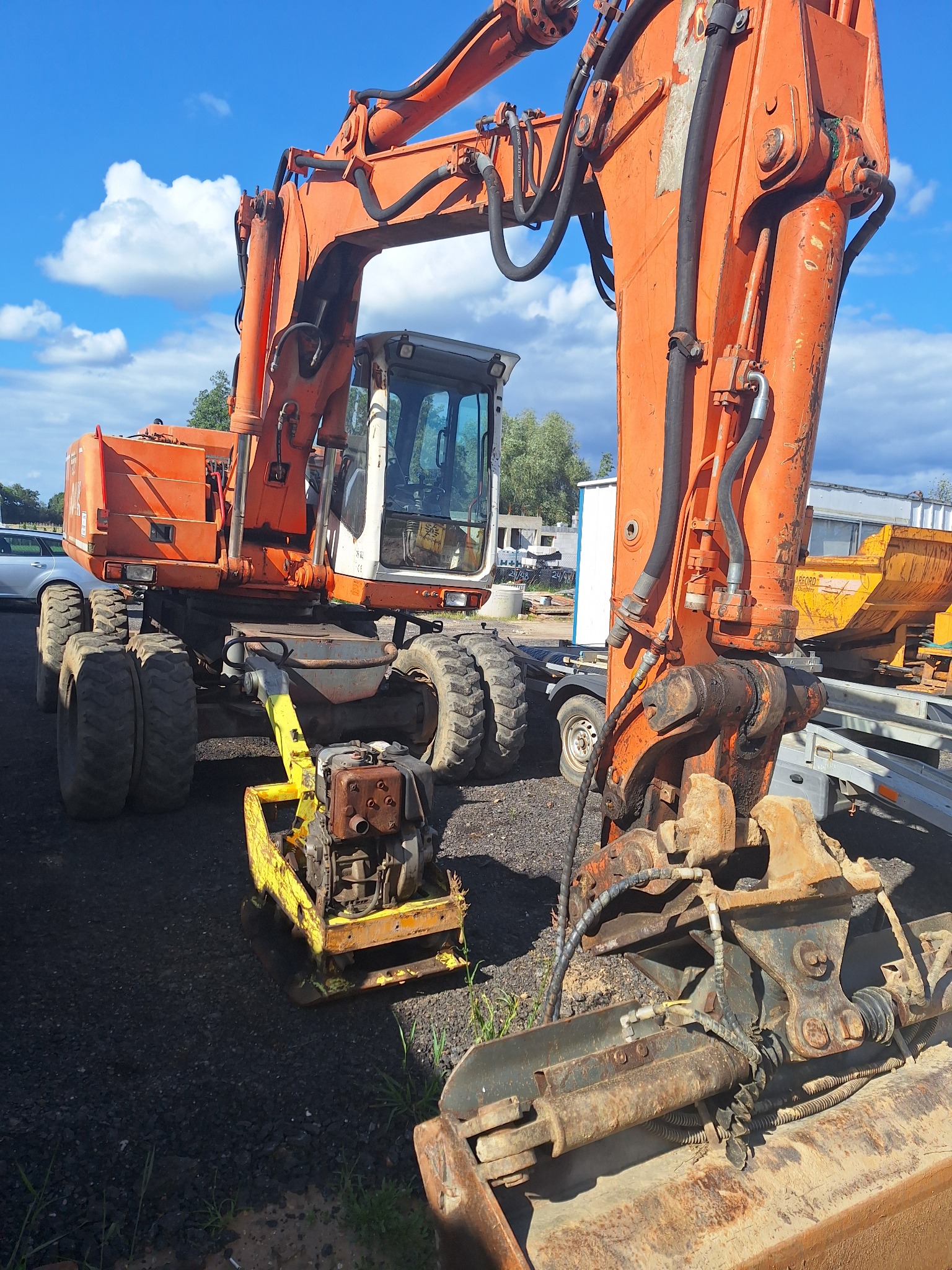 Pomarańczowa koparka kołowa z żółtą zagęszczarką w tle, na placu budowy, pod błękitnym niebem z chmurami.