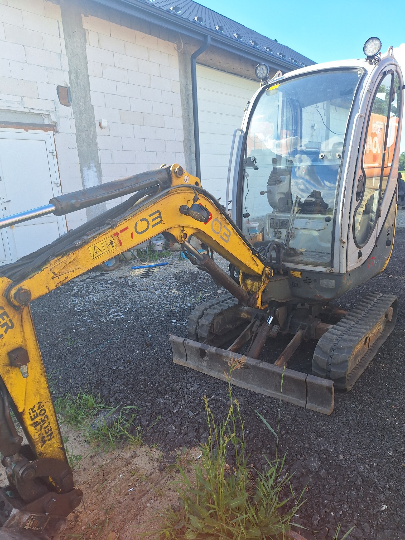 Żółta mini koparka Wacker Neuson na czarnym żwirze, obok ściany z białego betonu komórkowego i białych drzwi.