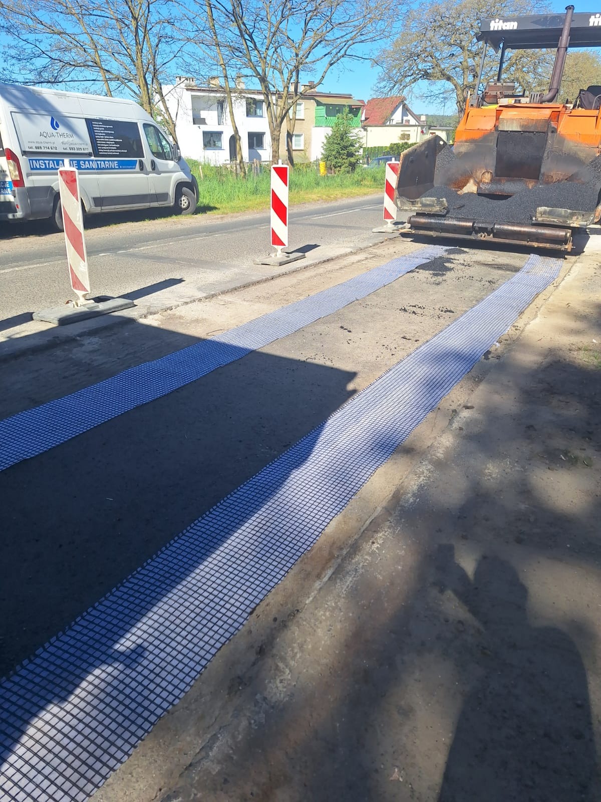Maszyna do układania asfaltu Titan na drodze w Skokowej, przygotowana podłoże z siatką wzmacniającą, widoczny samochód dostawczy z instalacjami sanitarnymi w tle.
