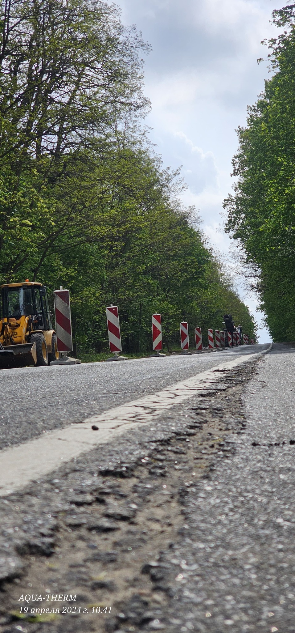 Uszkodzona asfaltowa droga w trakcie naprawy, widoczne ustawione pionowo biało-czerwone słupki ostrzegawcze, żółta koparka i zieleń drzew w tle.