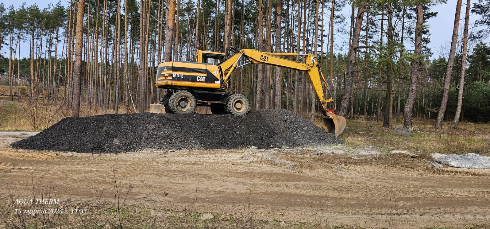 Żółta koparka CAT M312 na szczycie pryzmy ciemnej ziemi na tle lasu sosnowego.