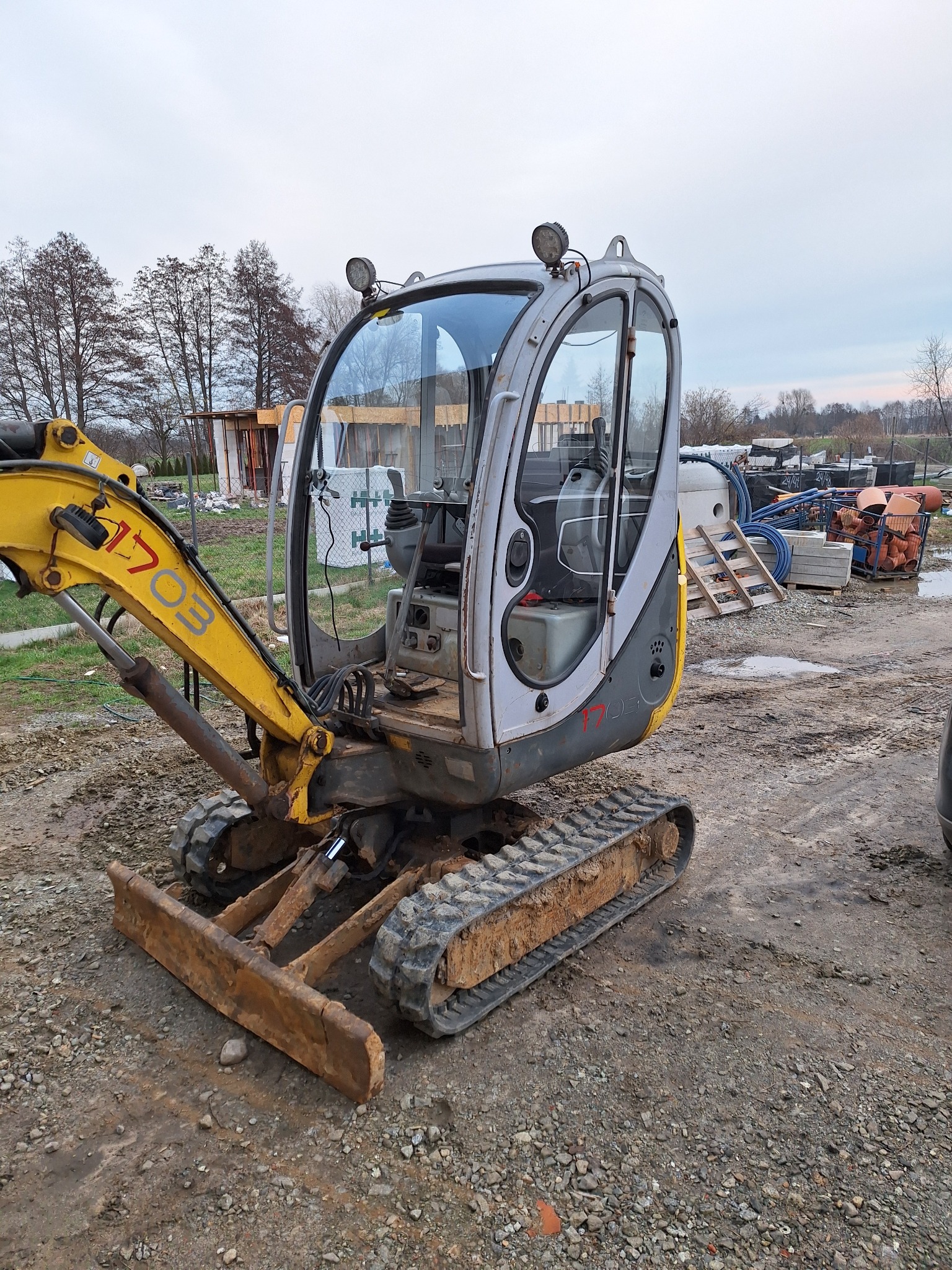 Mini koparka Wacker Neuson 1703, widoczne ślady użytkowania i zabrudzenia, na tle placu budowy z materiałami budowlanymi.