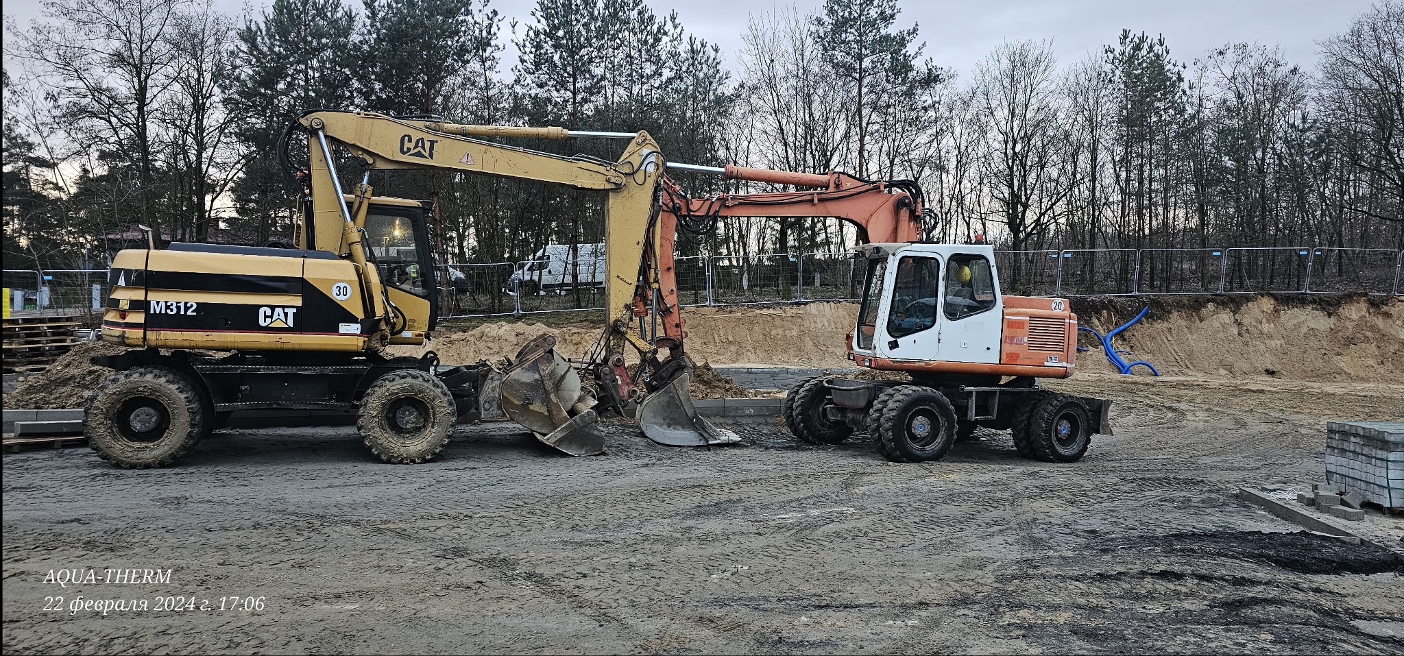 Dwa kołowe, żółto-czarny i pomarańczowo-biały,  koparki obok siebie na placu budowy z piaskiem i drzewami w tle.