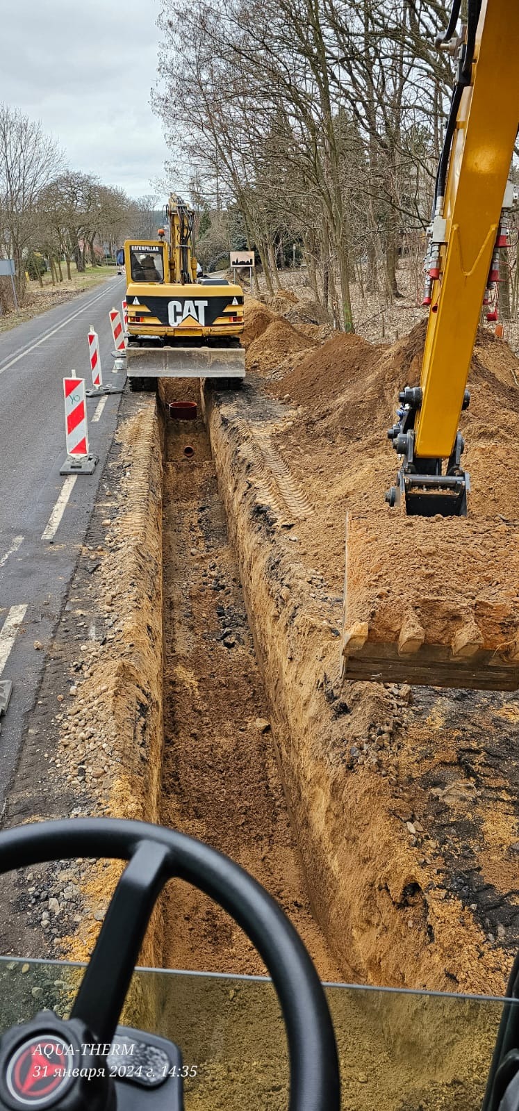 Żółta koparka CAT podczas wykonywania wykopu liniowego obok asfaltowej drogi, widok z kabiny operatora, z przodu widoczna kierownica, w tle drzewa bez liści i oznakowanie drogowe.