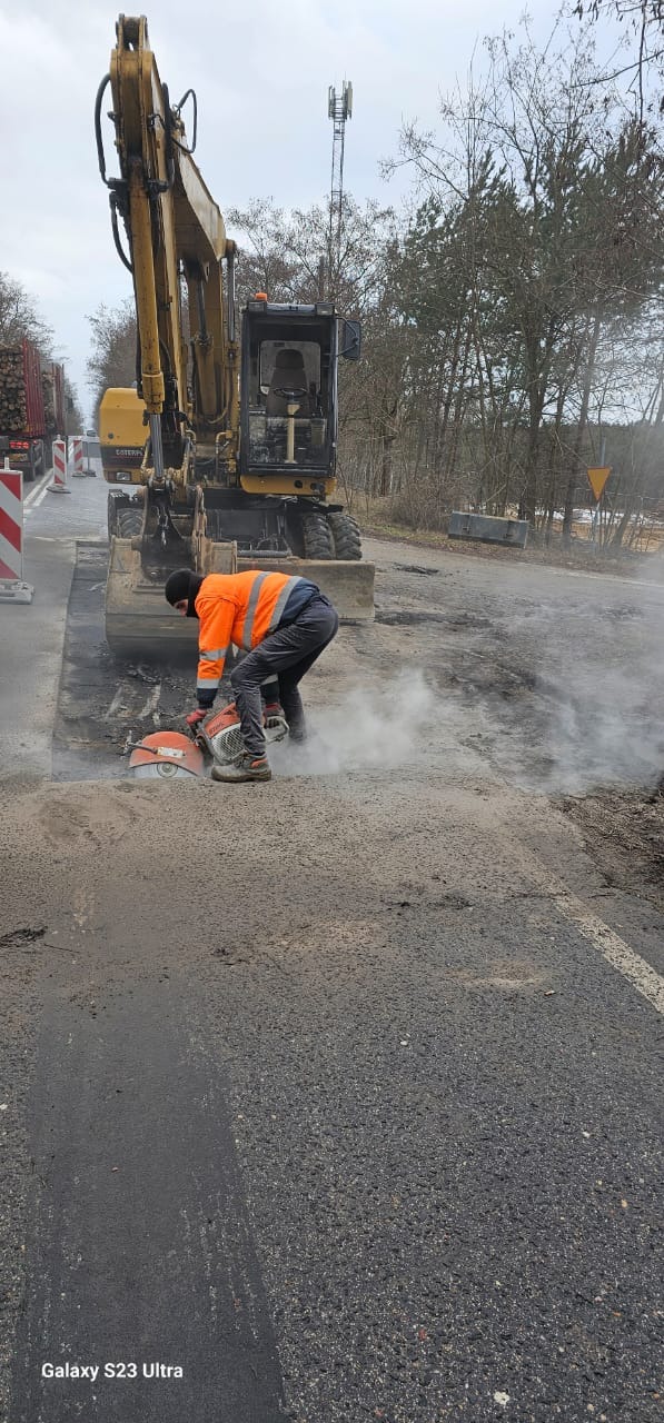 Pracownik w pomarańczowej kamizelce tnie asfaltową nawierzchnię drogi za pomocą piły spalinowej, obok stoi żółta koparka, w tle drzewa i maszt telekomunikacyjny.