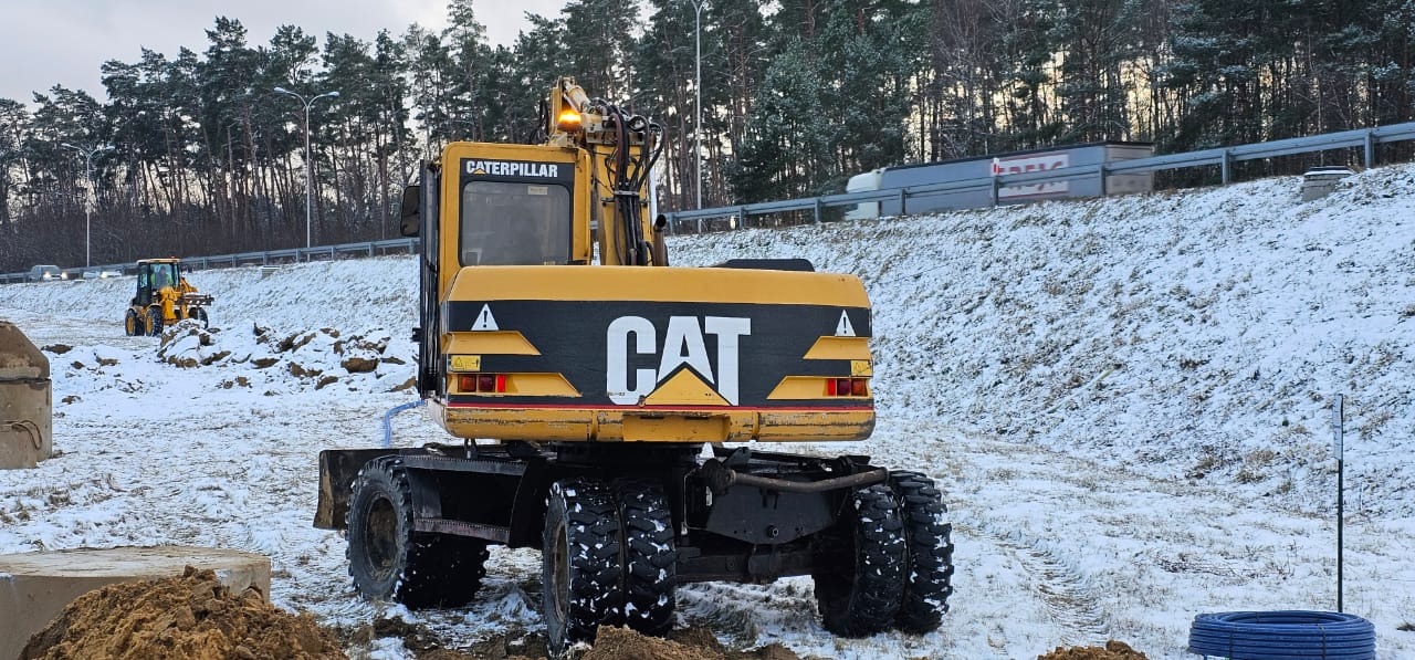 Żółta koparka kołowa CAT na zasypanym śniegiem placu budowy, w tle droga ekspresowa i las.