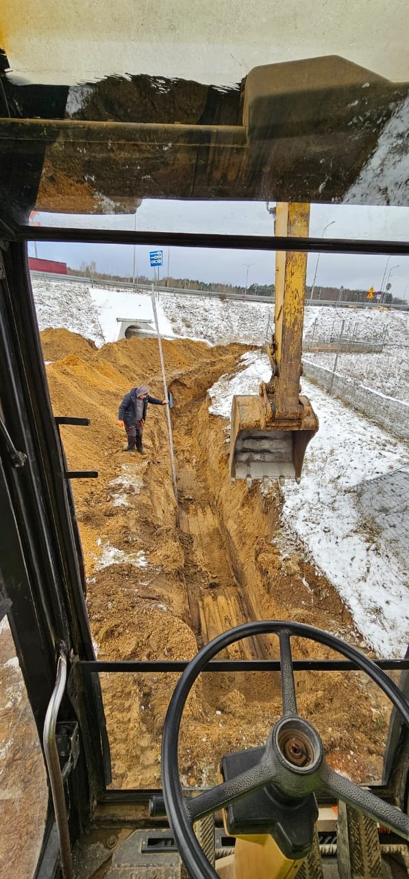 Widok z kabiny koparki na wykopany rów w ziemi, pracownik mierzy głębokość, w tle ośnieżone pobocze drogi.