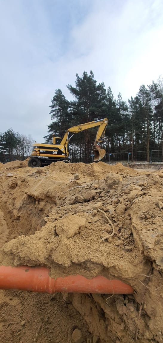 Żółta koparka CAT M312 na wzgórzu z piasku, w tle drzewa i pochmurne niebo, na pierwszym planie pomarańczowa rura w wykopie.