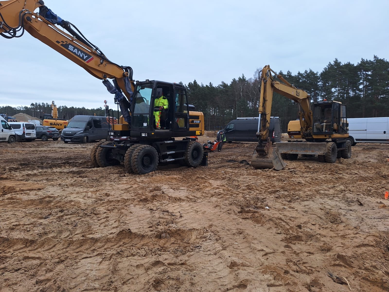 Dwa żółte koparki kołowe SANY i CAT na placu budowy, teren przygotowany, widoczne ślady opon i łyżki, w tle las i samochody dostawcze.