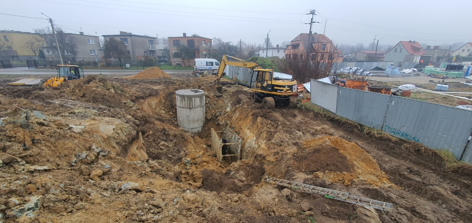 Żółta koparka CAT przy wykopie z betonowym kręgiem w ziemi, widoczne zabezpieczenia wykopu, drabina, w tle budynki mieszkalne i pochmurne niebo.