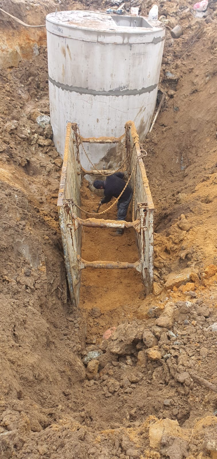 Pracownik z łopatą w wykopie zabezpieczonym metalową szalunką, obok betonowy zbiornik szamba.
