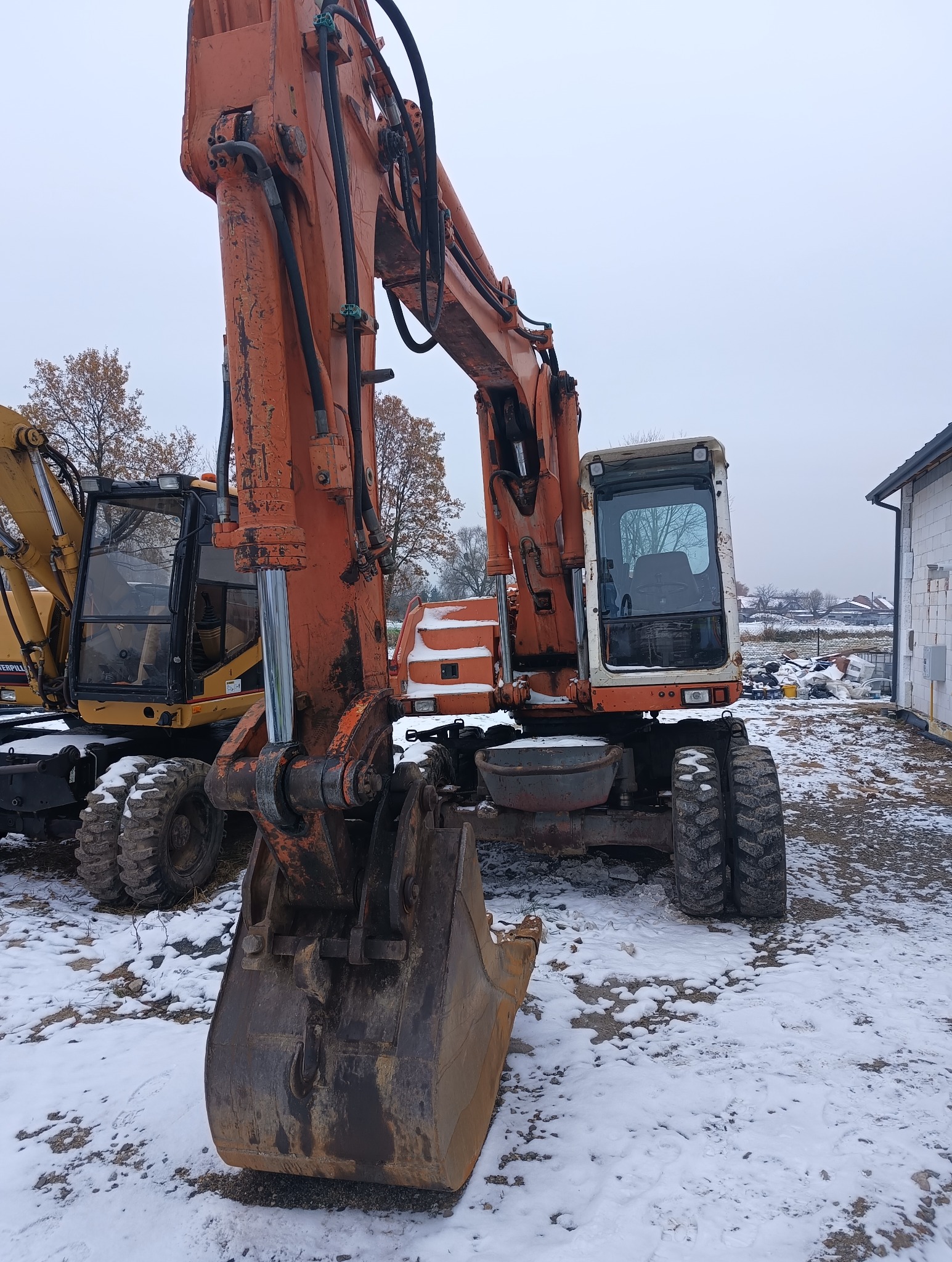 Pomarańczowa koparka kołowa z łyżką na zaśnieżonym placu budowy, obok żółta koparka, w tle budynek w stanie surowym i drzewa bez liści.