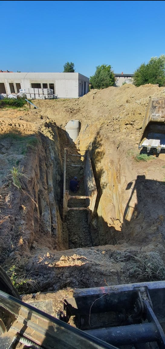Głęboki wykop w ziemi zabezpieczony drewnianą konstrukcją, pracownik w dole, widoczny fragment koparki, w tle budynek w trakcie budowy.