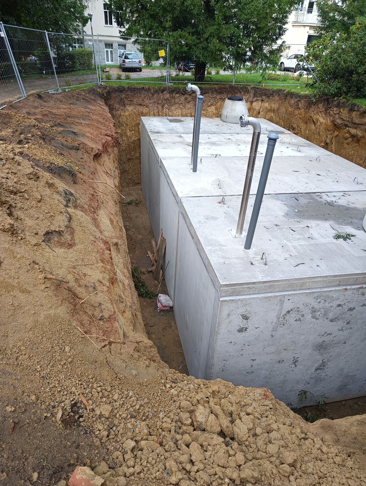 Betonowy zbiornik w wykopie, widoczne wystające rury i ziemia, w tle ogrodzenie i budynki.