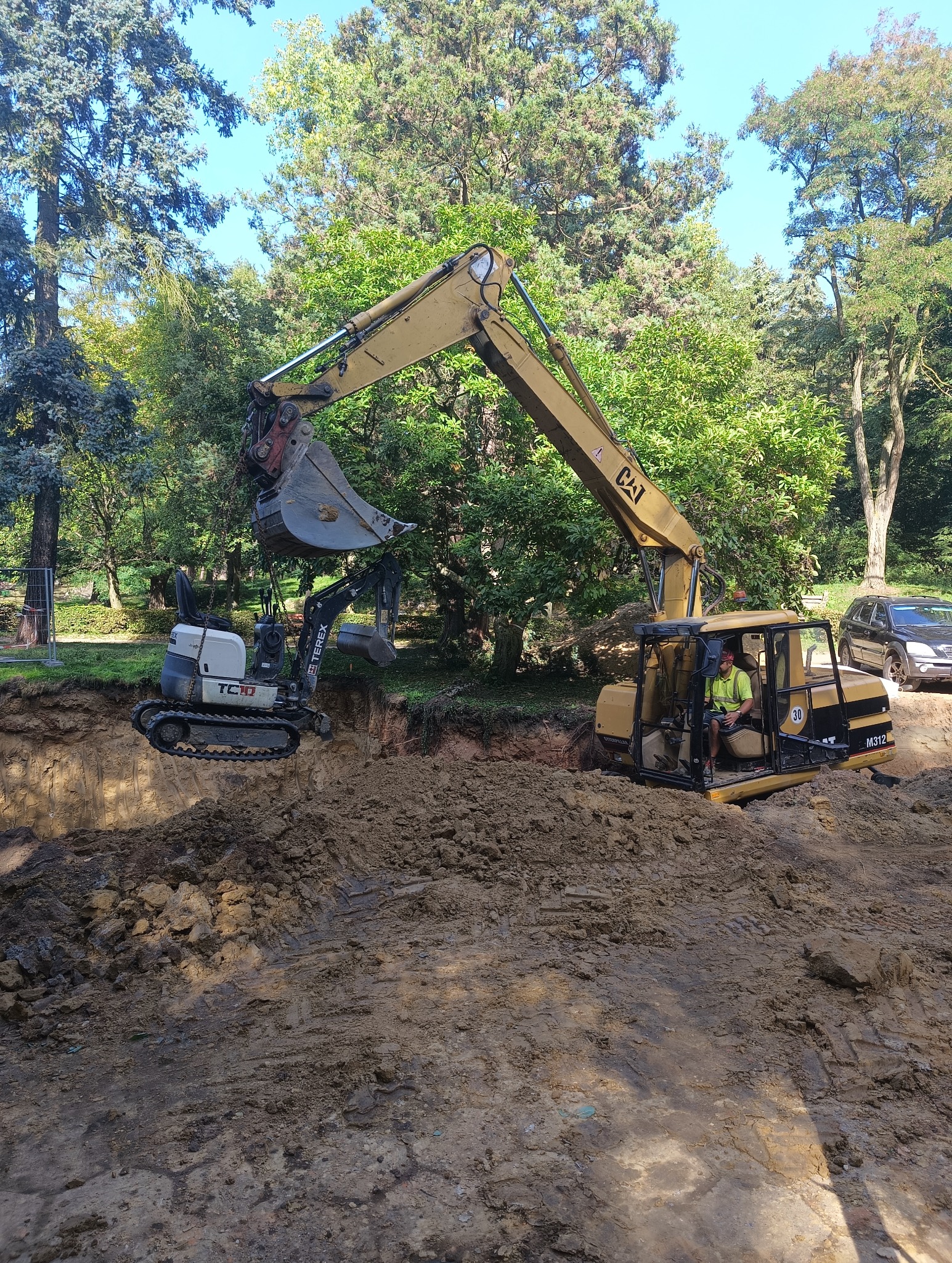Żółta koparka CAT M312 z operatorem w kabinie, obok mniejsza koparka Terex TC16, obie pracują w wykopie na tle drzew i zaparkowanego samochodu.