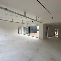 TipTopDom.pl - Wnętrze domu w stanie deweloperskim ze szkieletem stalowym pod sufitem, widoczne duże okna przesuwne i wylewka betonowa na podłodze.