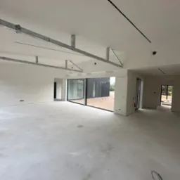 Wnętrze domu w stanie deweloperskim ze szkieletem stalowym pod sufitem, widoczne duże okna przesuwne i wylewka betonowa na podłodze.