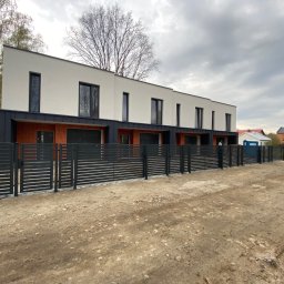 TipTopDom.pl - Szeregowe budynki jednorodzinne w stanie deweloperskim z elewacją w kolorze białym i czarnymi oknami, ogrodzone nowoczesnym, poziomym ogrodzeniem z metalowych sztachet na tle pochmurnego nieba.