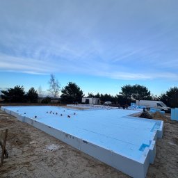 TipTopDom.pl - Ocieplona płyta fundamentowa z widocznymi elementami izolacji termicznej, na tle piaszczystego terenu budowy i częściowo zachmurzonego nieba.