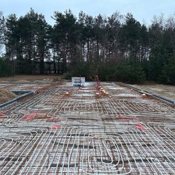 TipTopDom.pl - Zbrojenie ocieplanej płyty fundamentowej z systemem ogrzewania podłogowego, widoczna instalacja elektryczna i teren budowy z drzewami w tle.