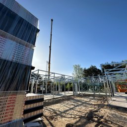 TipTopDom.pl - Konstrukcja domu na szkielecie stalowym w trakcie montażu, widoczne przygotowane pakiety płyt Balex Metal, żuraw budowlany w tle i podnośnik koszowy.