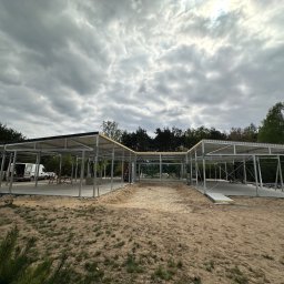 TipTopDom.pl - Konstrukcja szkieletowa budynku z zamontowanym dachem, widoczny van z otwartymi drzwiami w tle, teren piaszczysty, pochmurne niebo.