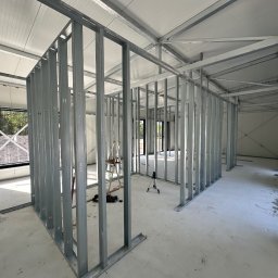 TipTopDom.pl - Wnętrze budynku w trakcie budowy, widoczne stalowe profile konstrukcyjne ścian działowych, poziomica laserowa na statywie, drabina i narzędzia na podłodze.