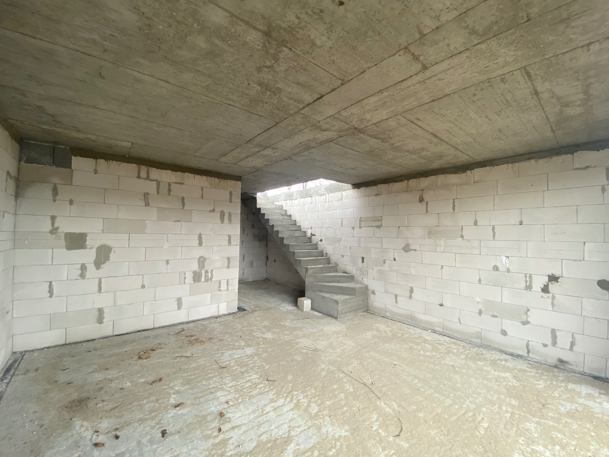 Wnętrze budynku w stanie surowym z betonu komórkowego, widoczne betonowe schody prowadzące na wyższą kondygnację, surowy betonowy strop.