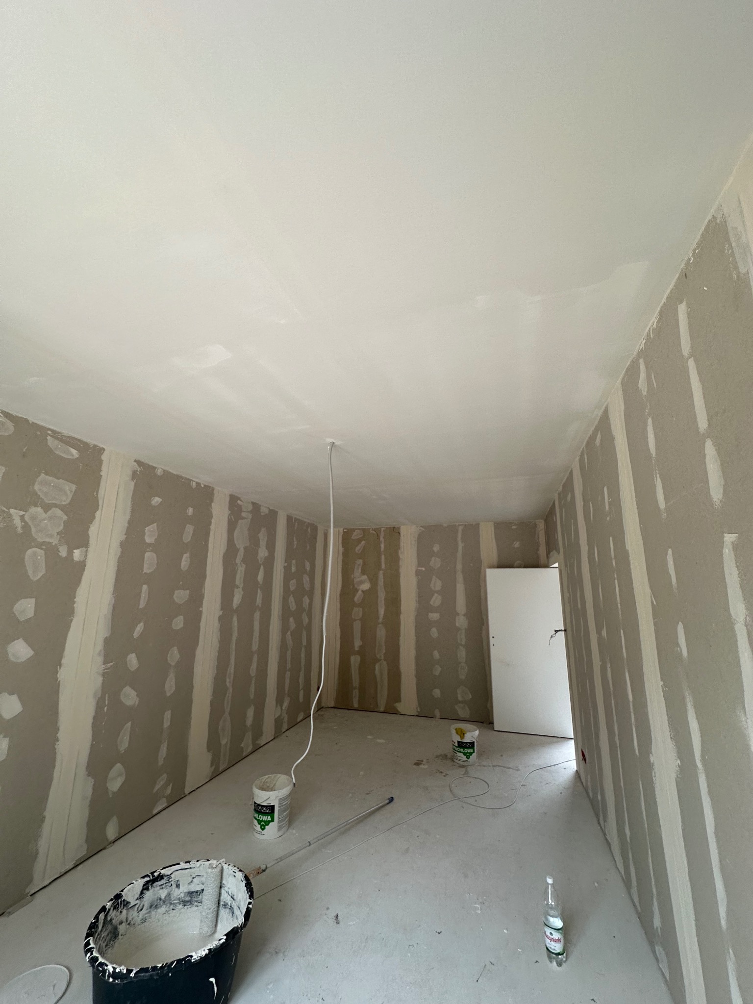 Wnętrze w trakcie prac wykończeniowych: ściany przygotowane do malowania, z widocznymi łączeniami płyt gipsowych, na podłodze wiadro z farbą i butelka wody mineralnej.