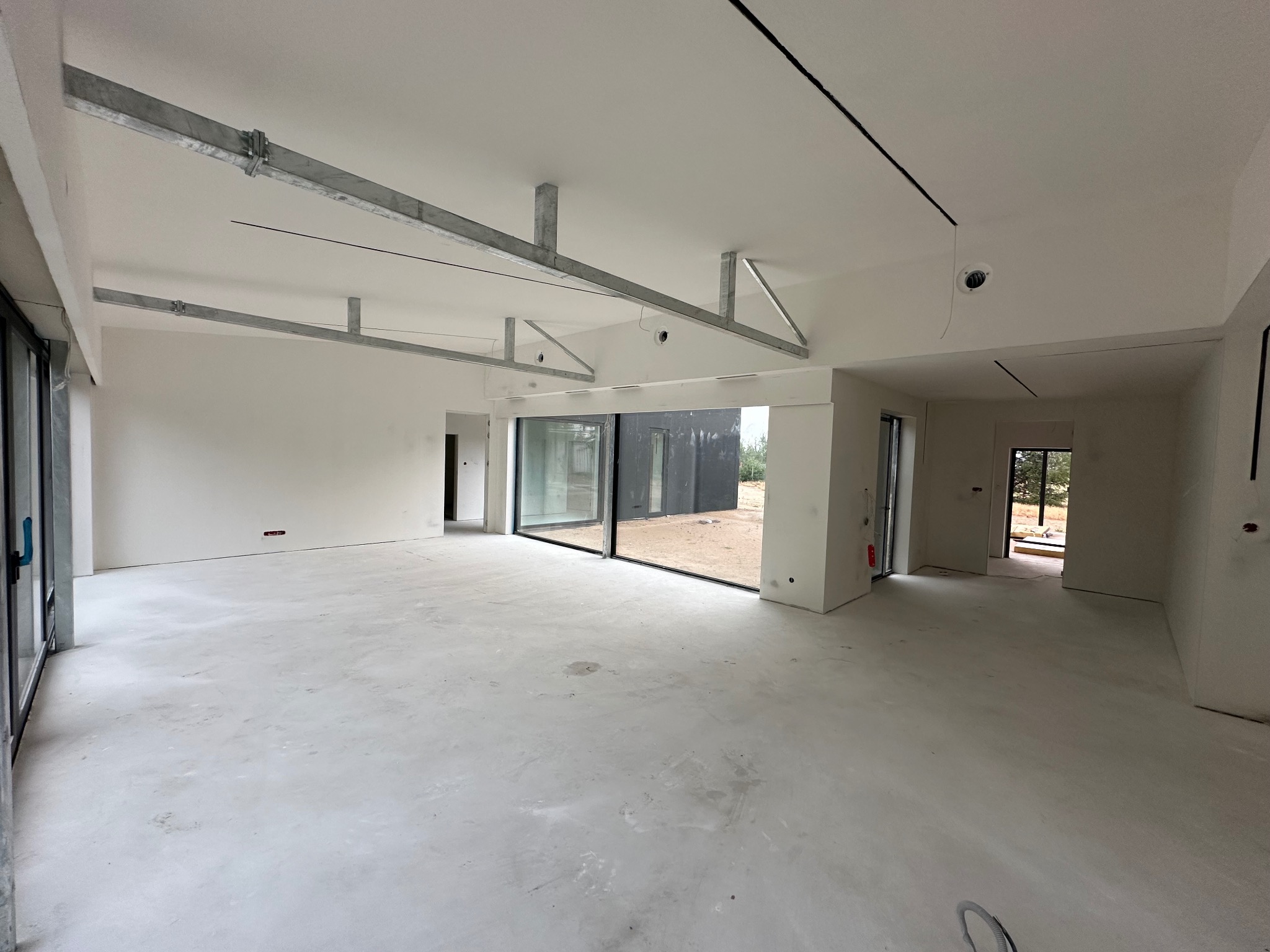 Wnętrze domu w stanie deweloperskim ze szkieletem stalowym pod sufitem, widoczne duże okna przesuwne i wylewka betonowa na podłodze.