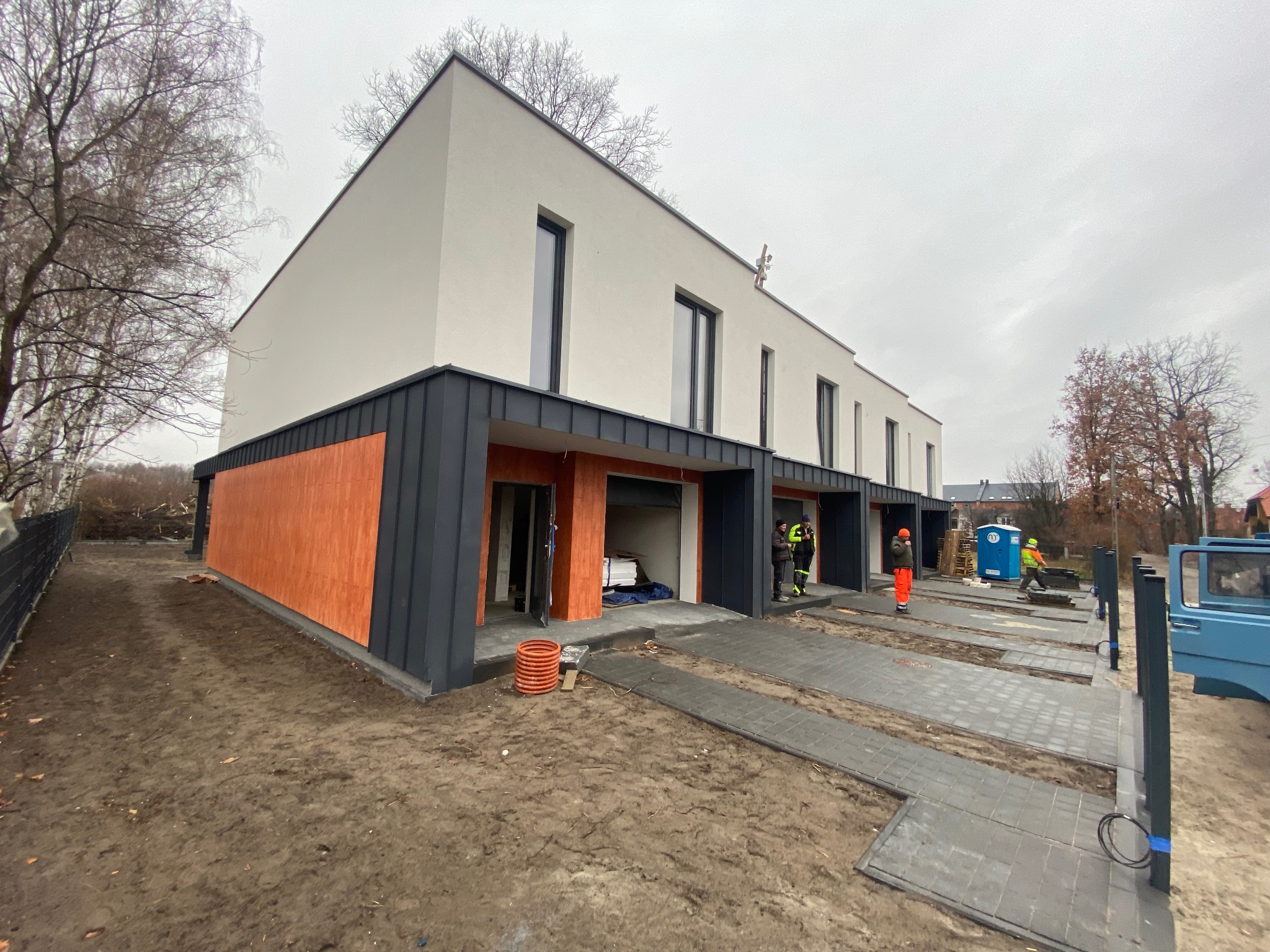 Szeregowy budynek jednorodzinny w stanie deweloperskim z elewacją w kolorach bieli, szarości i drewna, widoczni pracownicy w strojach roboczych oraz częściowo ułożona kostka brukowa na podjeździe.